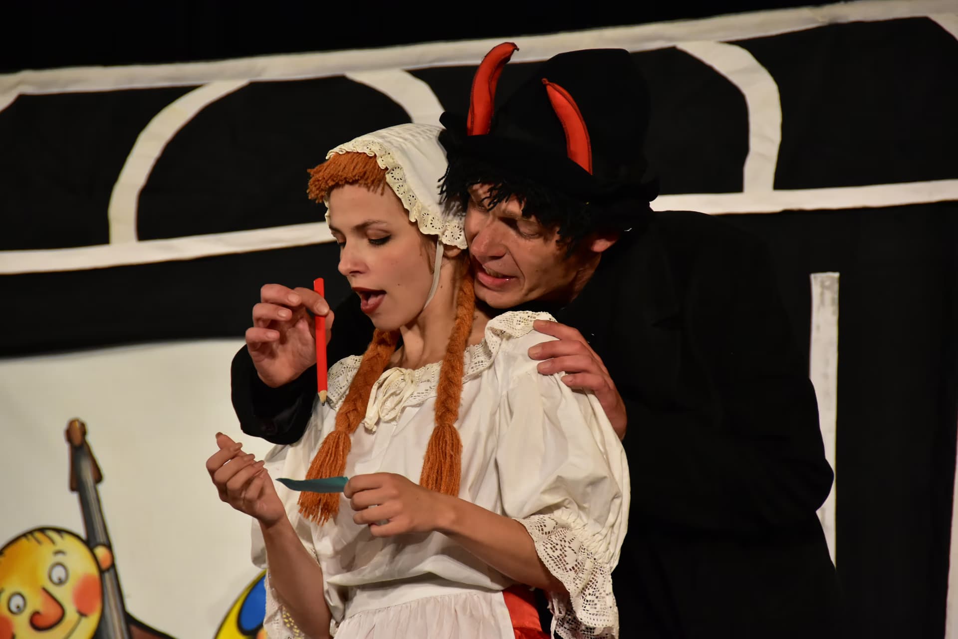 Aktor w kostiumie diabła obejmuje kobietę w stroju ludowym na scenie teatralnej.