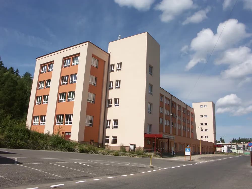 Moderní vícepatrová administrativní či školní budova stojí na zpevněné ploše pod modrou oblohou.