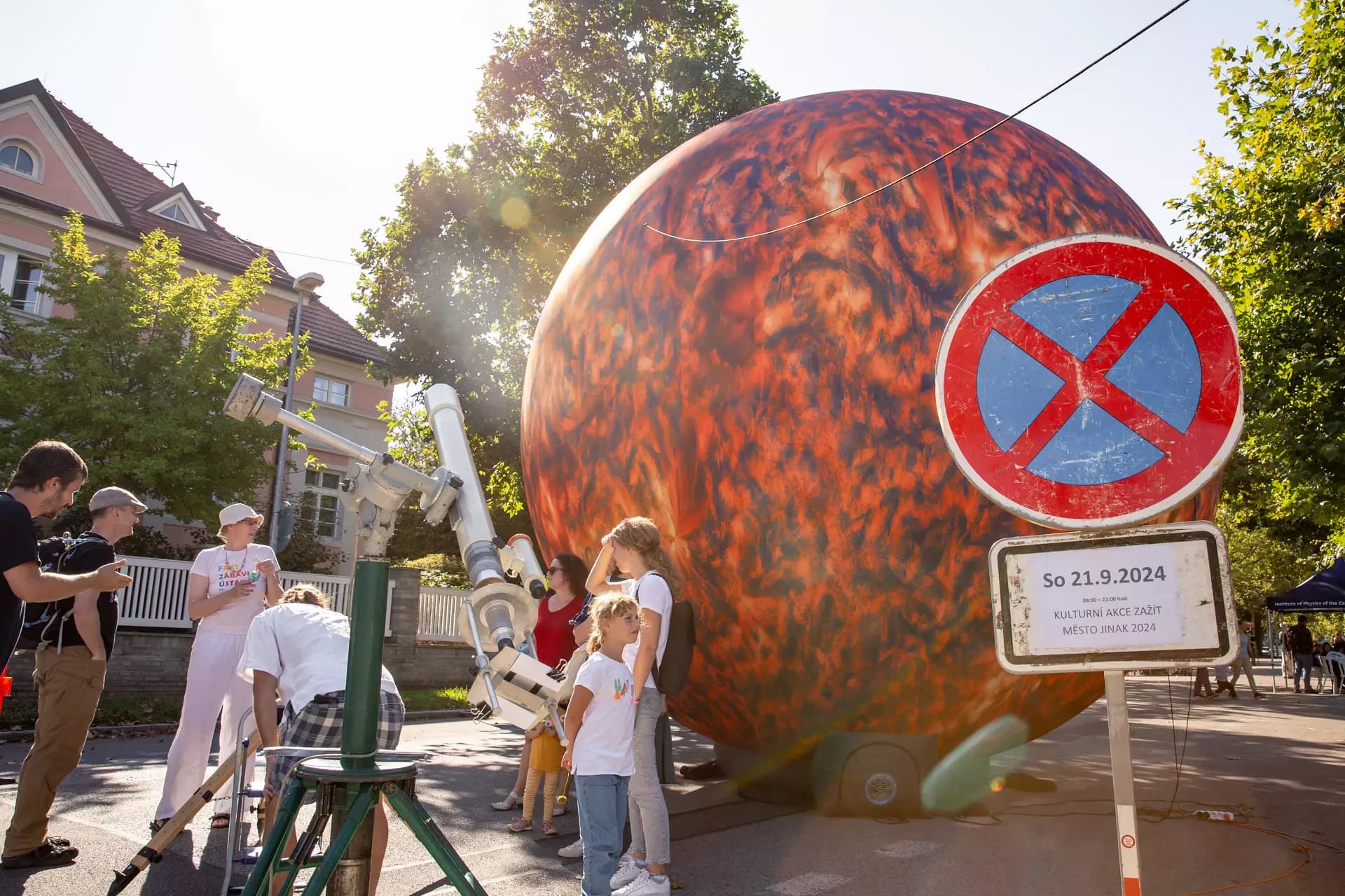 People observe a large inflatable sun model in a city square with a telescope nearby.