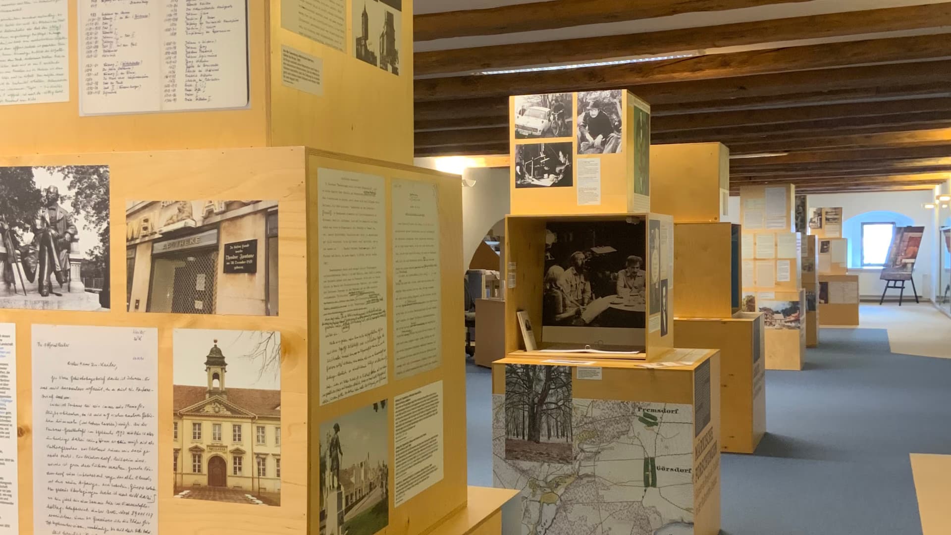 Dřevěné výstavní panely ve světlé místnosti obsahují četné historické fotografie a listy s tištěným textem.