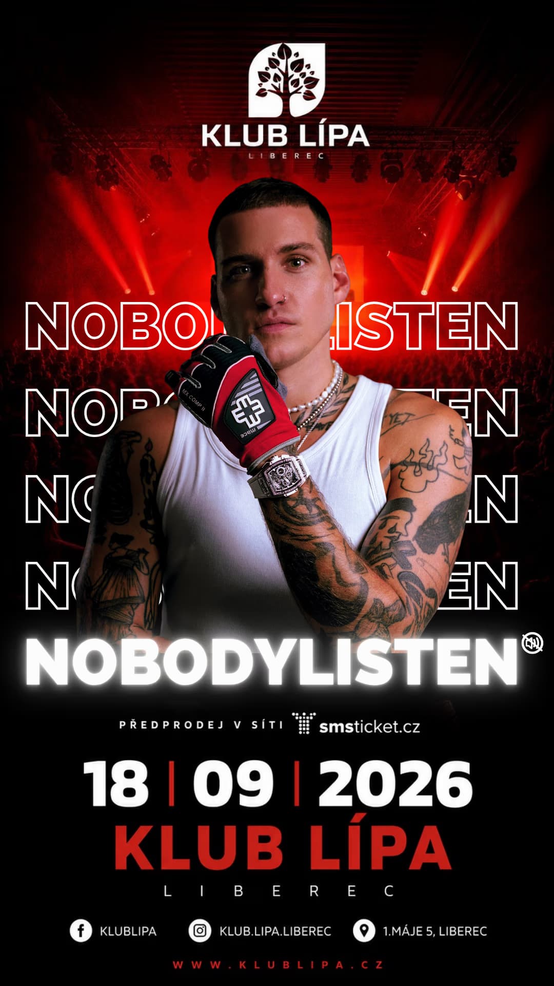 A tattooed man in a white tank top poses against a dark, red-lit concert stage background.
