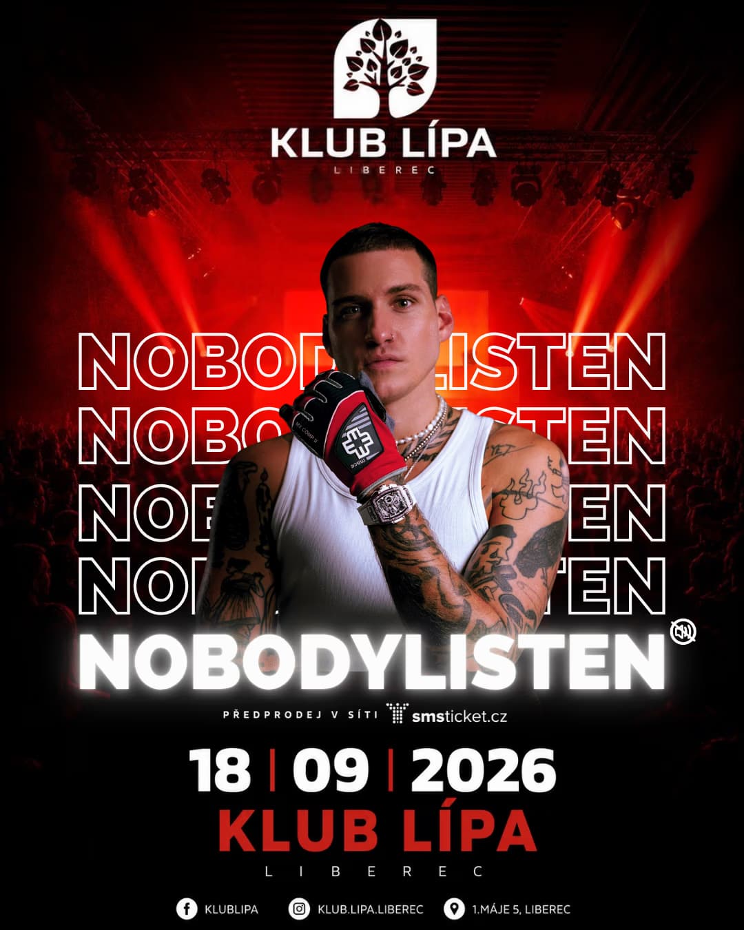 Tattooed man in a white tank top poses with a glove before a dark concert stage with red lights.