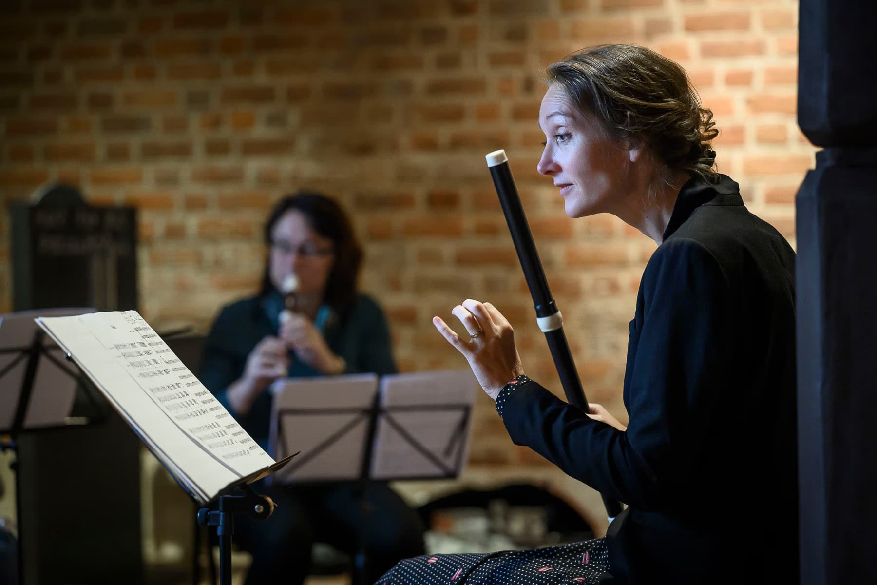 A female musician plays a wooden recorder in front of a brick wall during a concert.