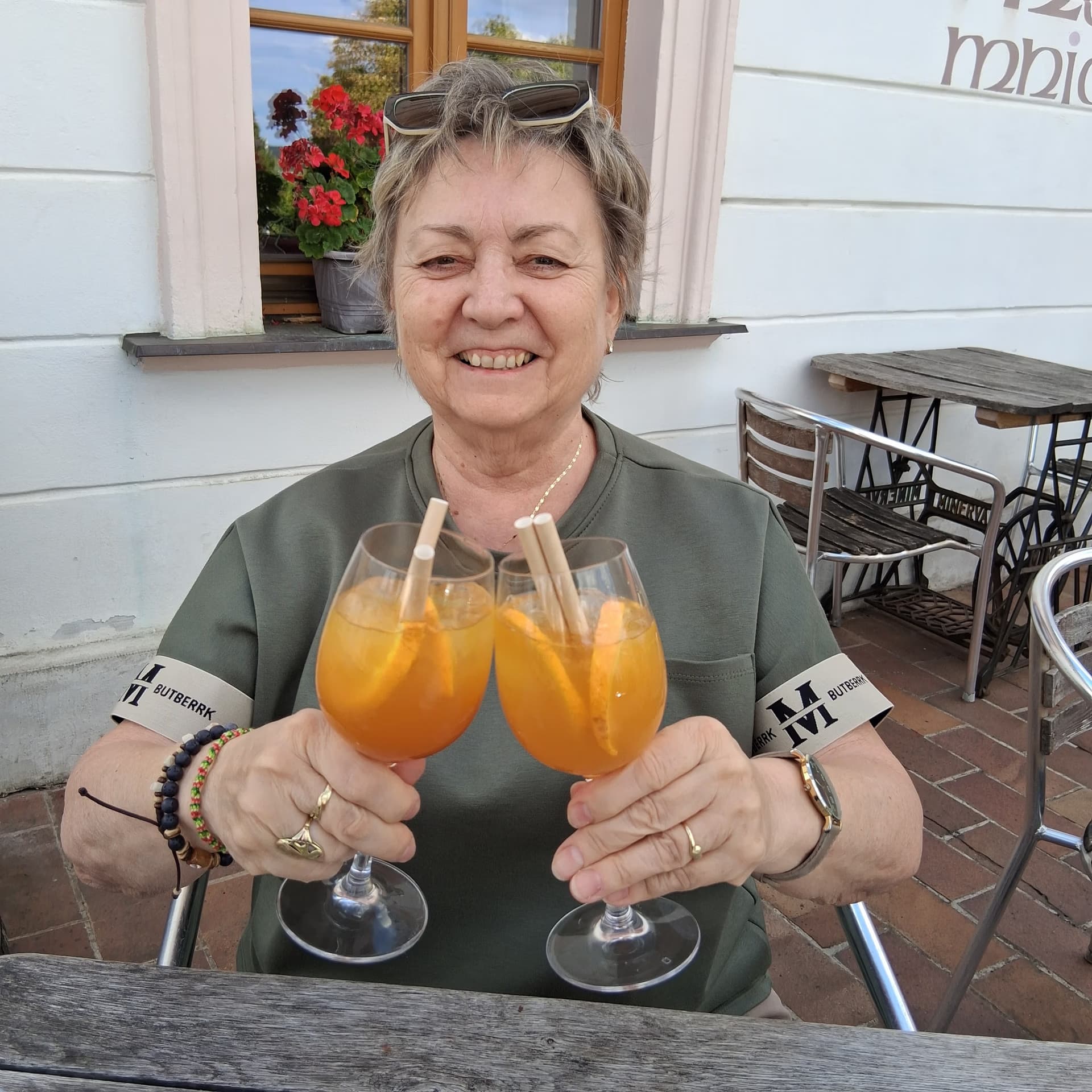 Uśmiechnięta kobieta trzyma dwa kieliszki z pomarańczowym drinkiem na tarasie restauracji.