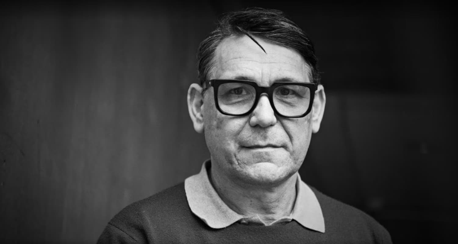Ein mittelalter Mann mit dunklem Haar und Brille vor einem dunklen Hintergrund.