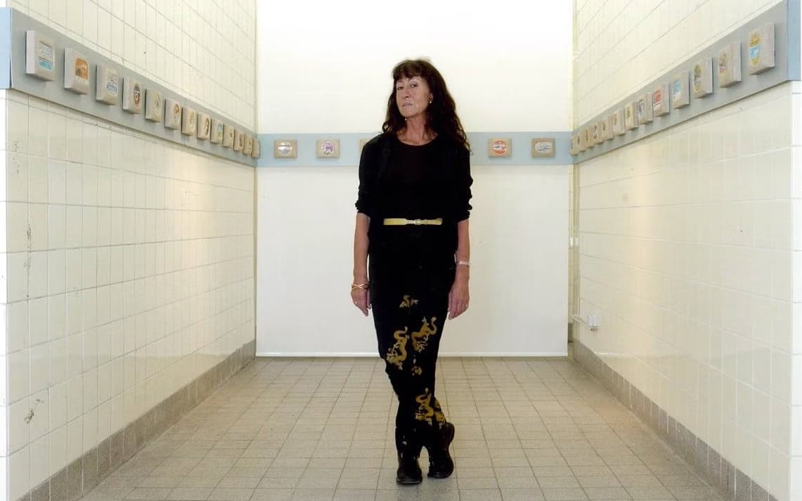 A woman stands in a narrow white tiled hallway decorated with rows of small framed artworks.