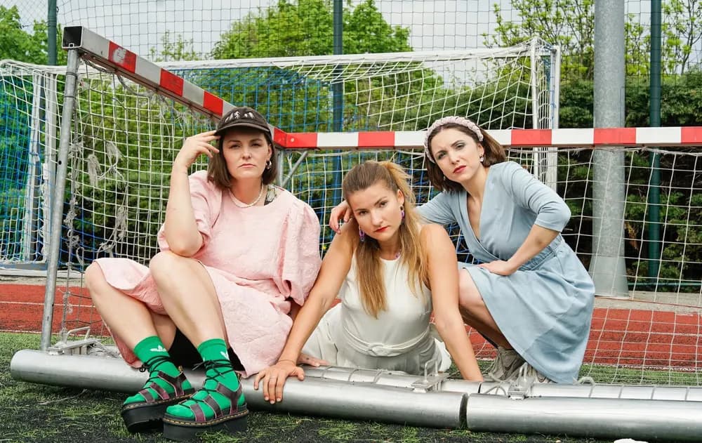 Three young women in dresses sit posing on a soccer goal frame at a sports field.