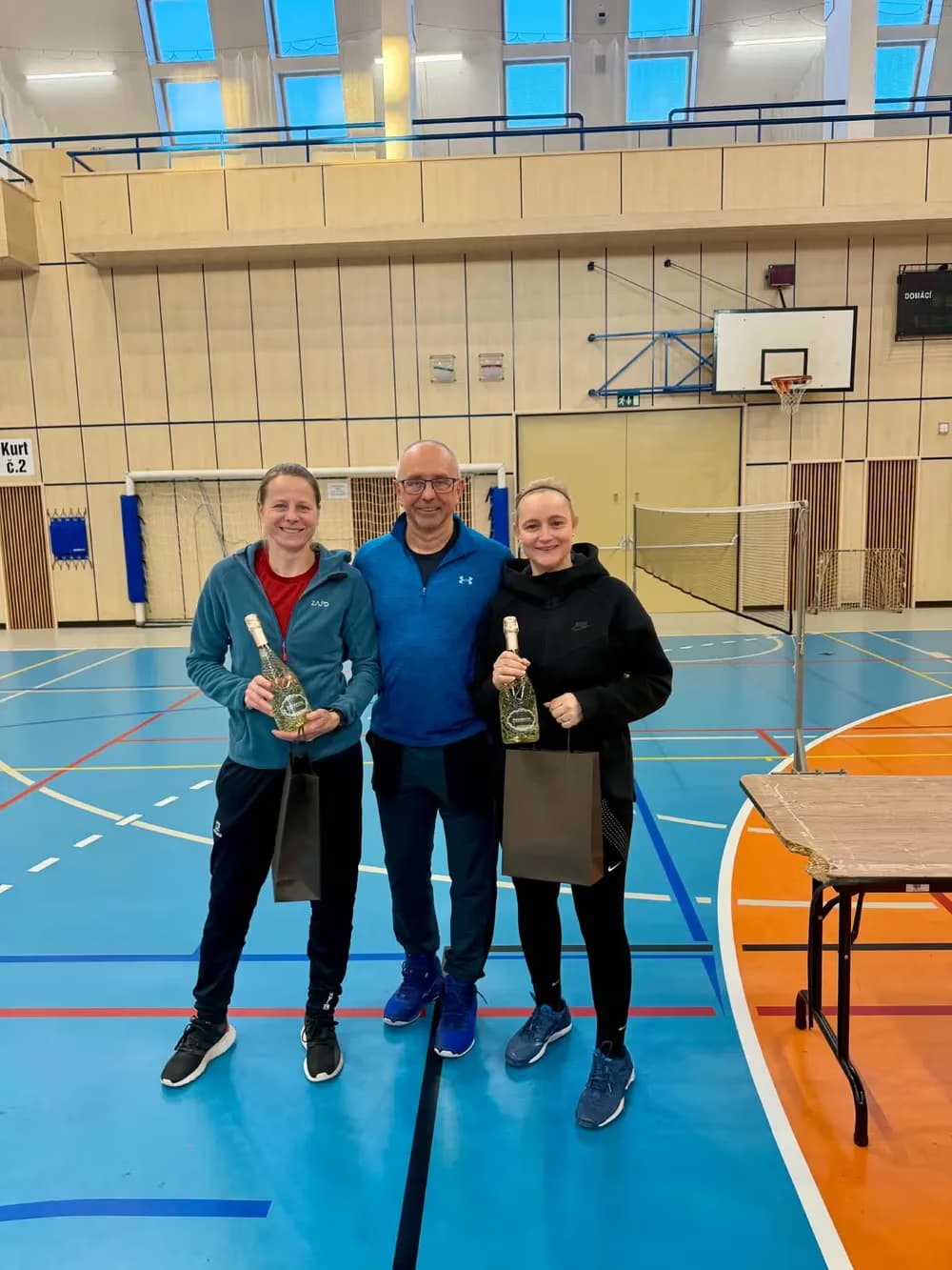 Tři lidé s lahvemi vína a taškami stojí v tělocvičně po skončení sportovního turnaje.