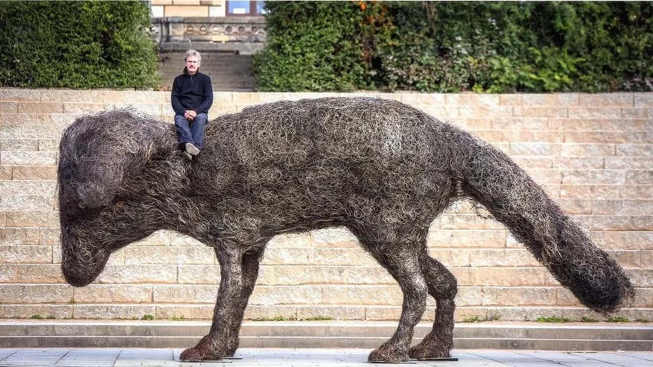Mężczyzna siedzi na dużej, fakturowanej rzeźbie lisa wykonanej z ciemnego, splątanego materiału organicznego.