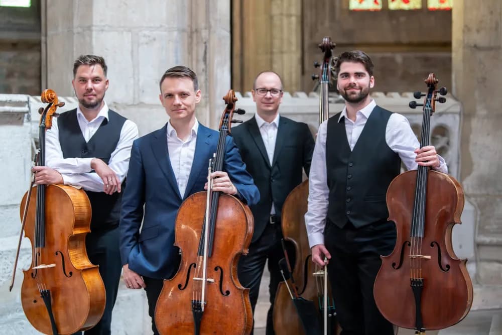 Four men in formal attire stand with cellos in a stone hall interior.