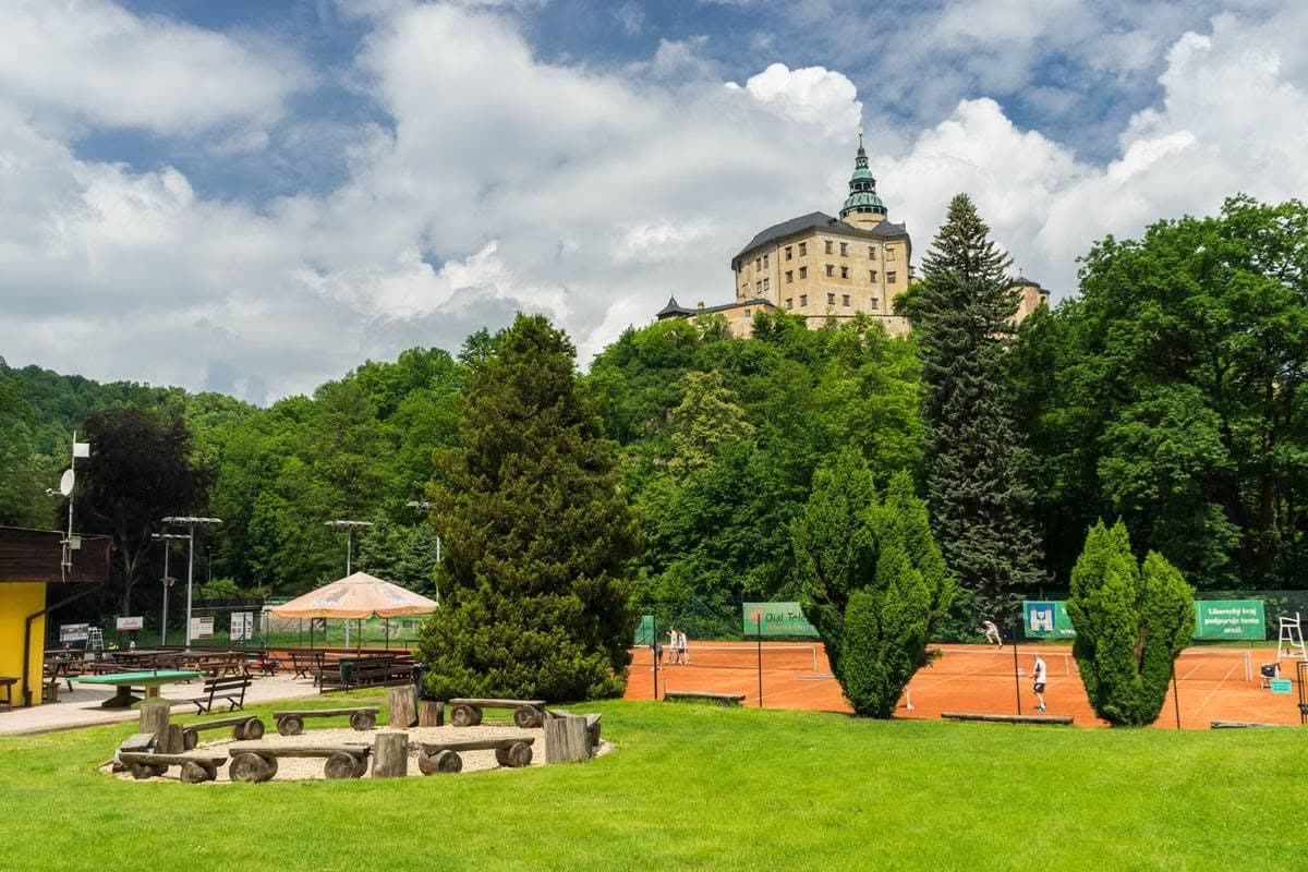 A clay tennis court situated below a castle perched on a hill, surrounded by green trees.