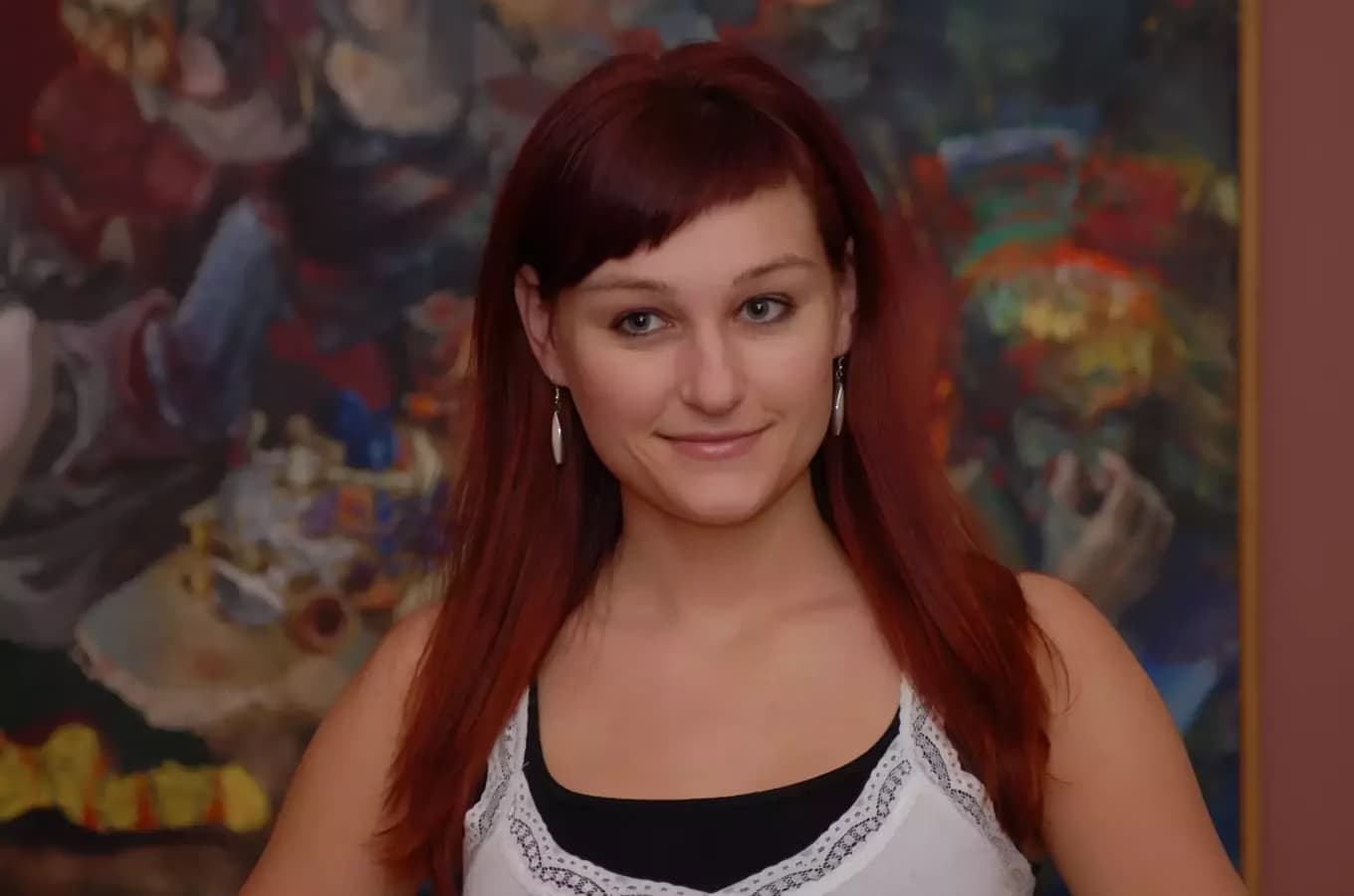A young woman with reddish hair smiling in front of a colorful abstract painting.