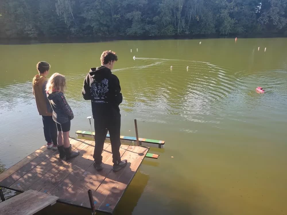 Tři lidé stojí na dřevěném molu a sledují malou dálkově ovládanou loď na rybníku.
