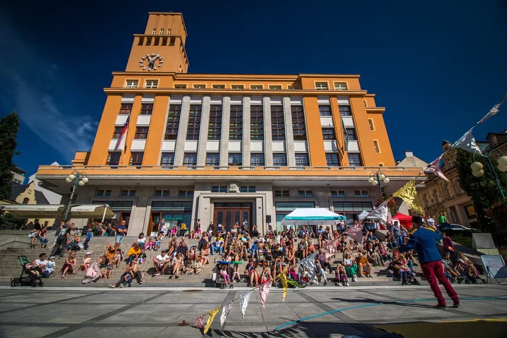 Crowds of people sit on the steps of a large ochre-colored town hall building on a sunny day.