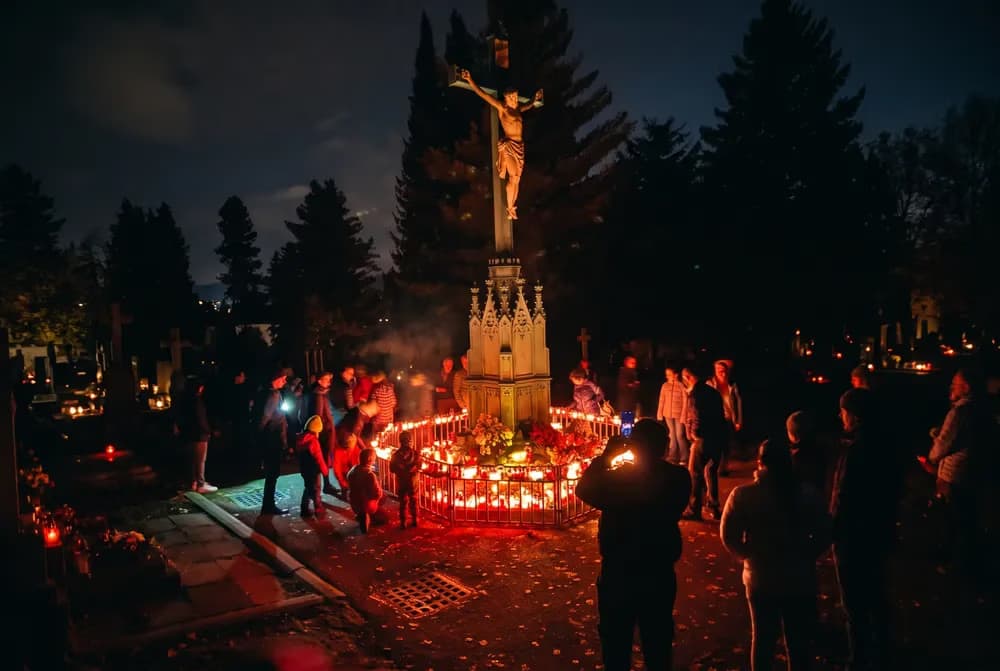 Lidé se večer shromažďují kolem osvětleného pomníku s křížem na hřbitově.