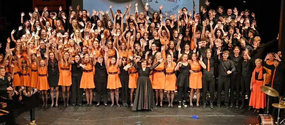 Ein großer Chor in schwarz-orangen Outfits winkt dem Publikum auf der Bühne zu.