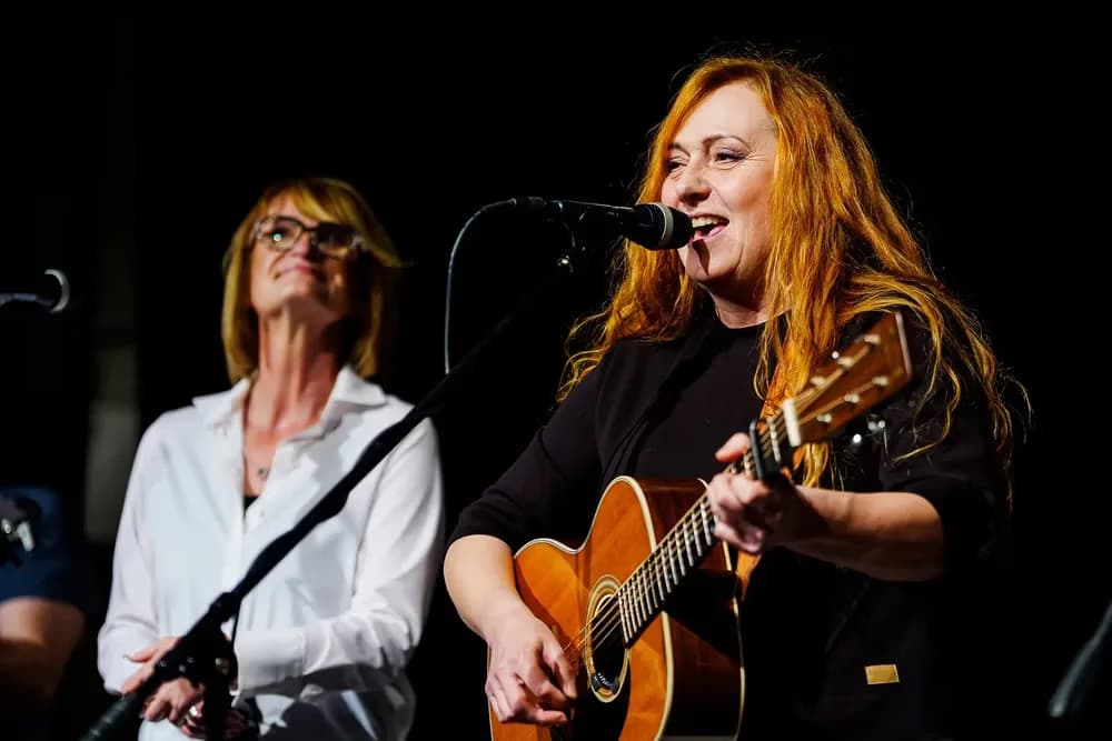 A woman with red hair plays an acoustic guitar and sings into a microphone on stage.