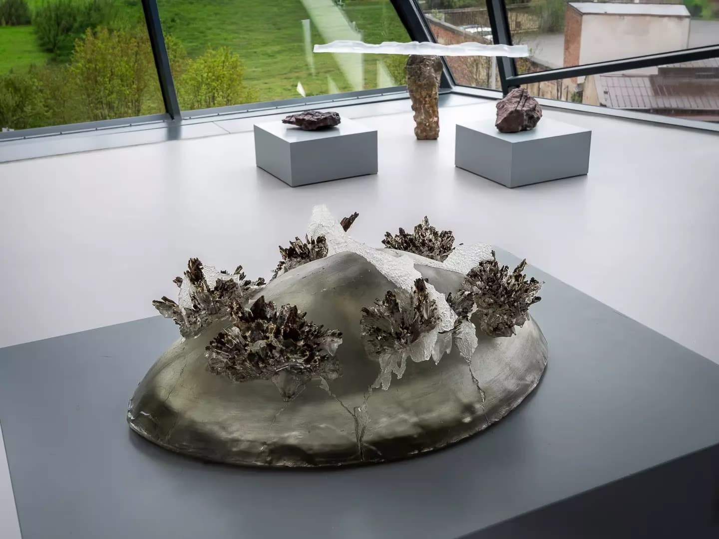 Artistic metal sculpture with crystal-like protrusions sits on a display stand in a museum room.