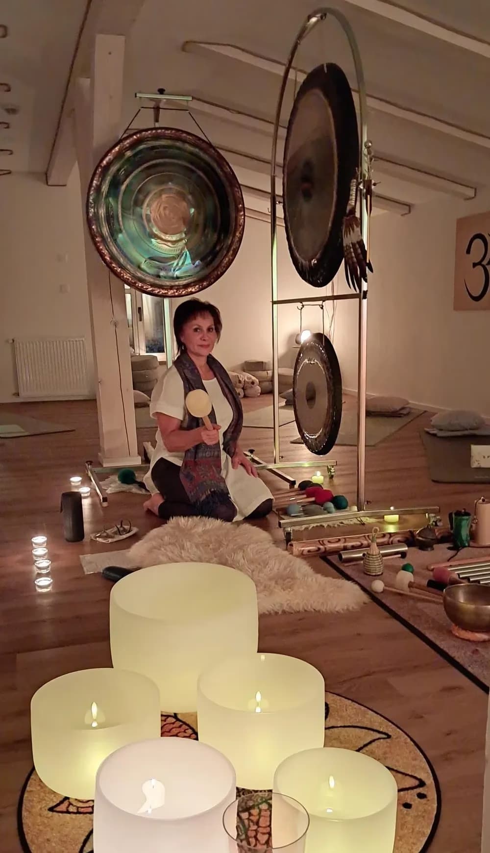 A woman kneels in a meditation studio surrounded by glowing singing bowls and large gongs.
