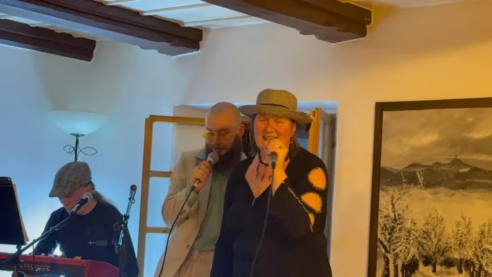 Ein Duo singt in Mikrofone, während ein Musiker im Hintergrund auf einem Keyboard spielt.