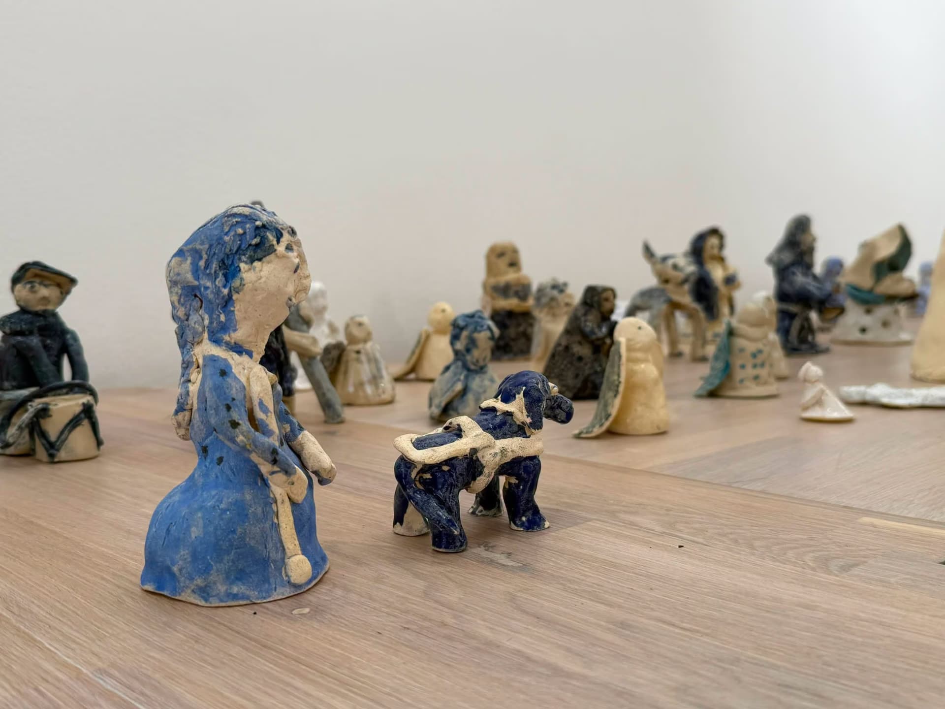 Many small, handmade clay figurines of people and animals arranged on a light wooden table.