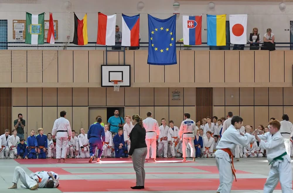 Zawodnicy judo w hali sportowej z międzynarodowymi flagami wiszącymi na balustradzie balkonu.