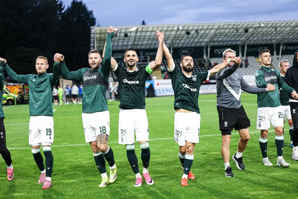 Fotbalisté v zelených dresech se drží za ruce a slaví na hřišti po zápase.