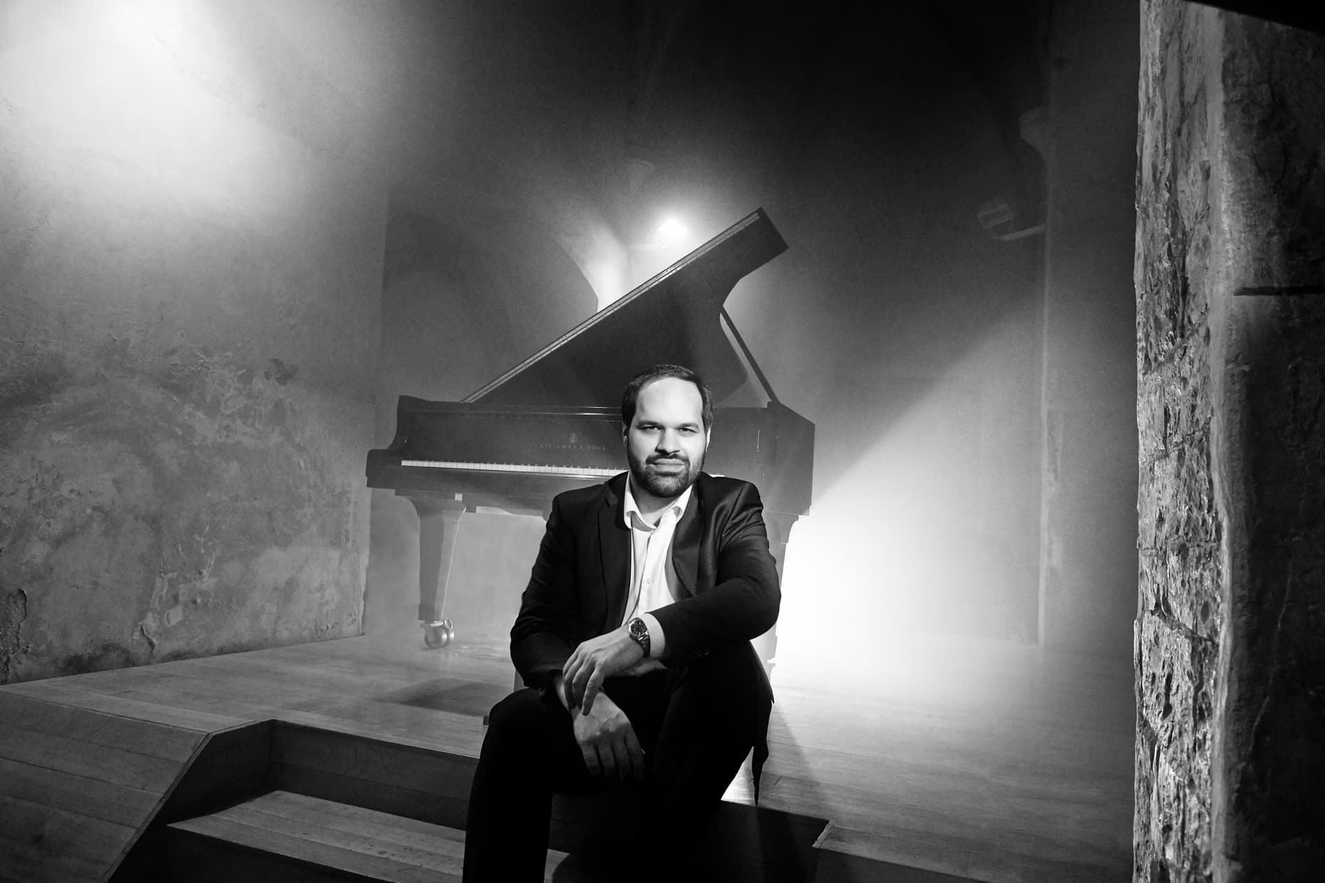 A bearded man in a suit sits on stairs before a grand piano in a dimly lit, atmospheric room.