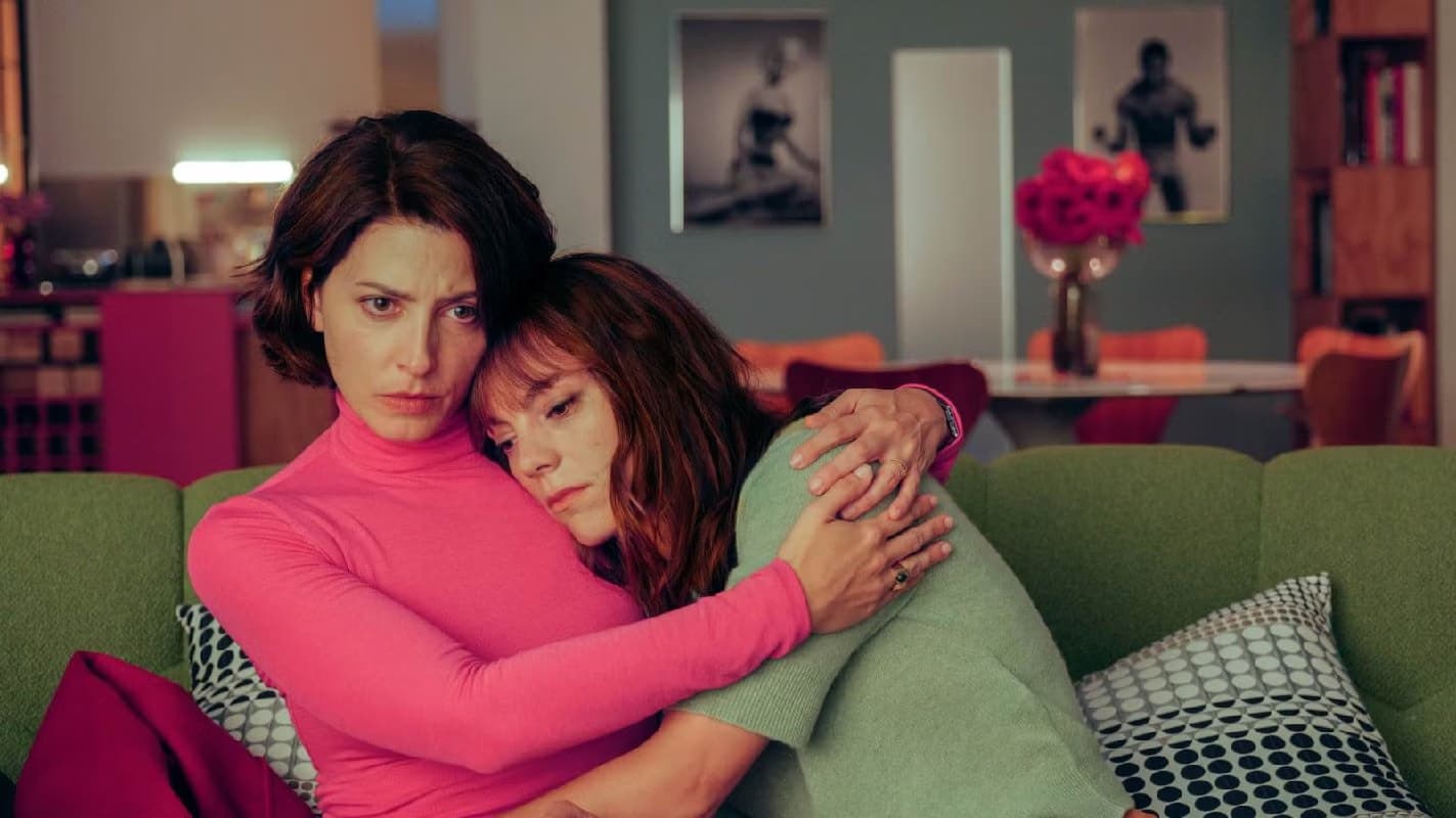 Two women sitting on a green sofa, one comforting the other in a domestic living room.