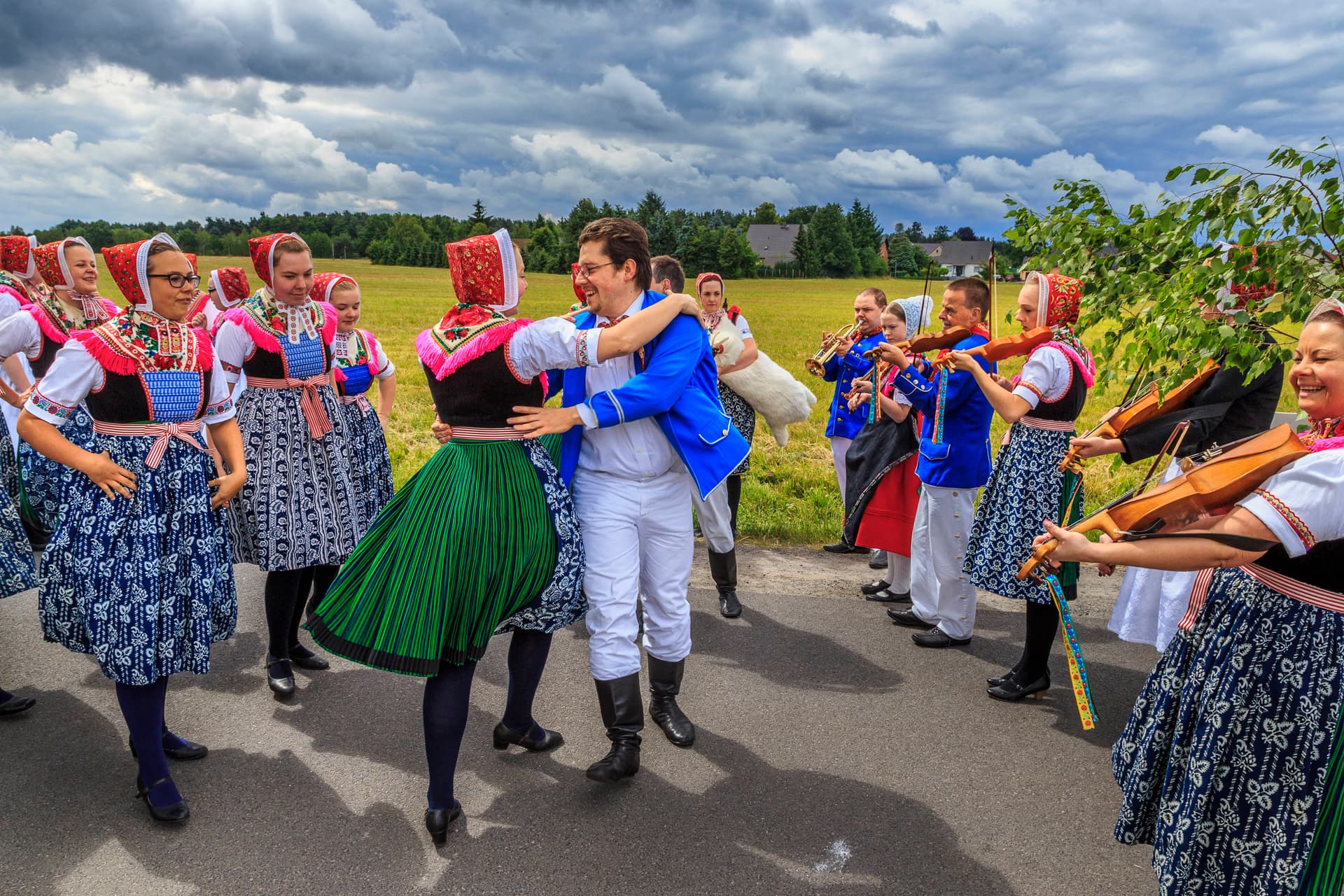 Ludzie w tradycyjnych strojach ludowych tańczą i grają na skrzypcach na ścieżce na zewnątrz.