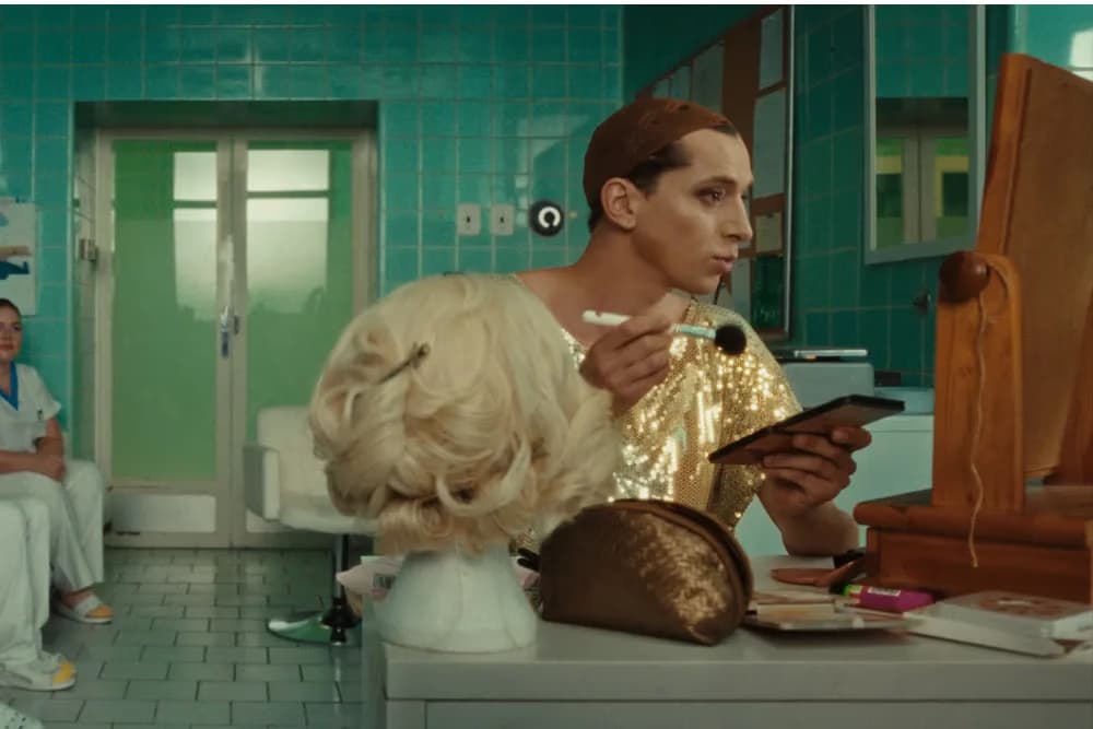 A person in a gold shirt applies makeup before a mirror in a tiled room with a blonde wig.
