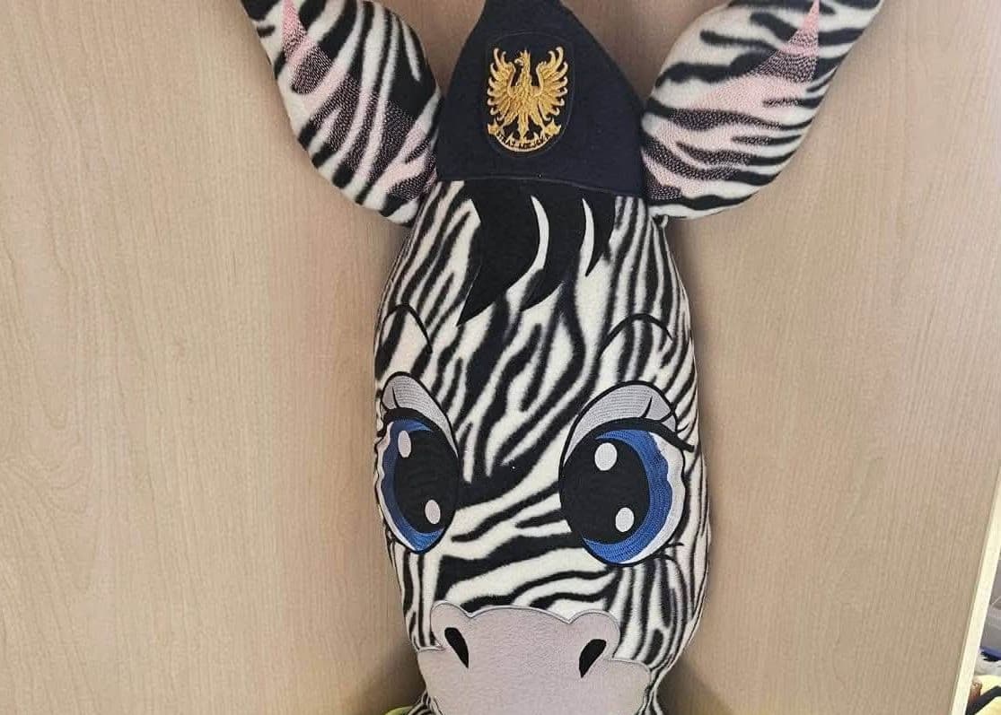 Plyšová hlava zebry s malou modrou čepicí se zlatým znakem na hlavě.