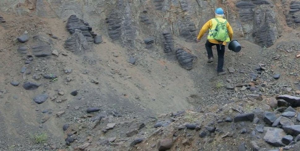 A person with a yellow jacket and blue helmet hikes uphill through a rocky, grey quarry.