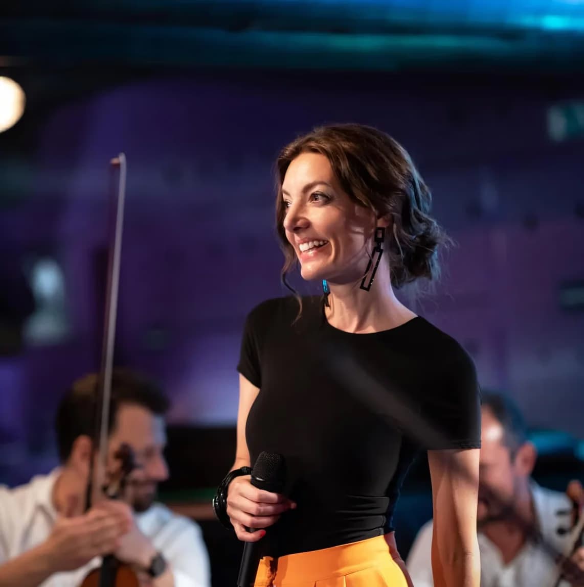 Smiling woman in a black top holding a microphone on stage during a musical performance.