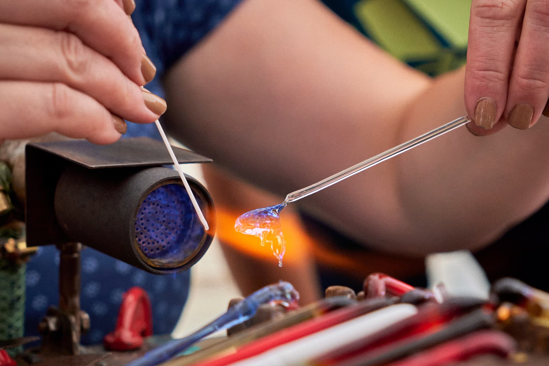 Close-up of hands melting a blue glass rod over a small burner flame.