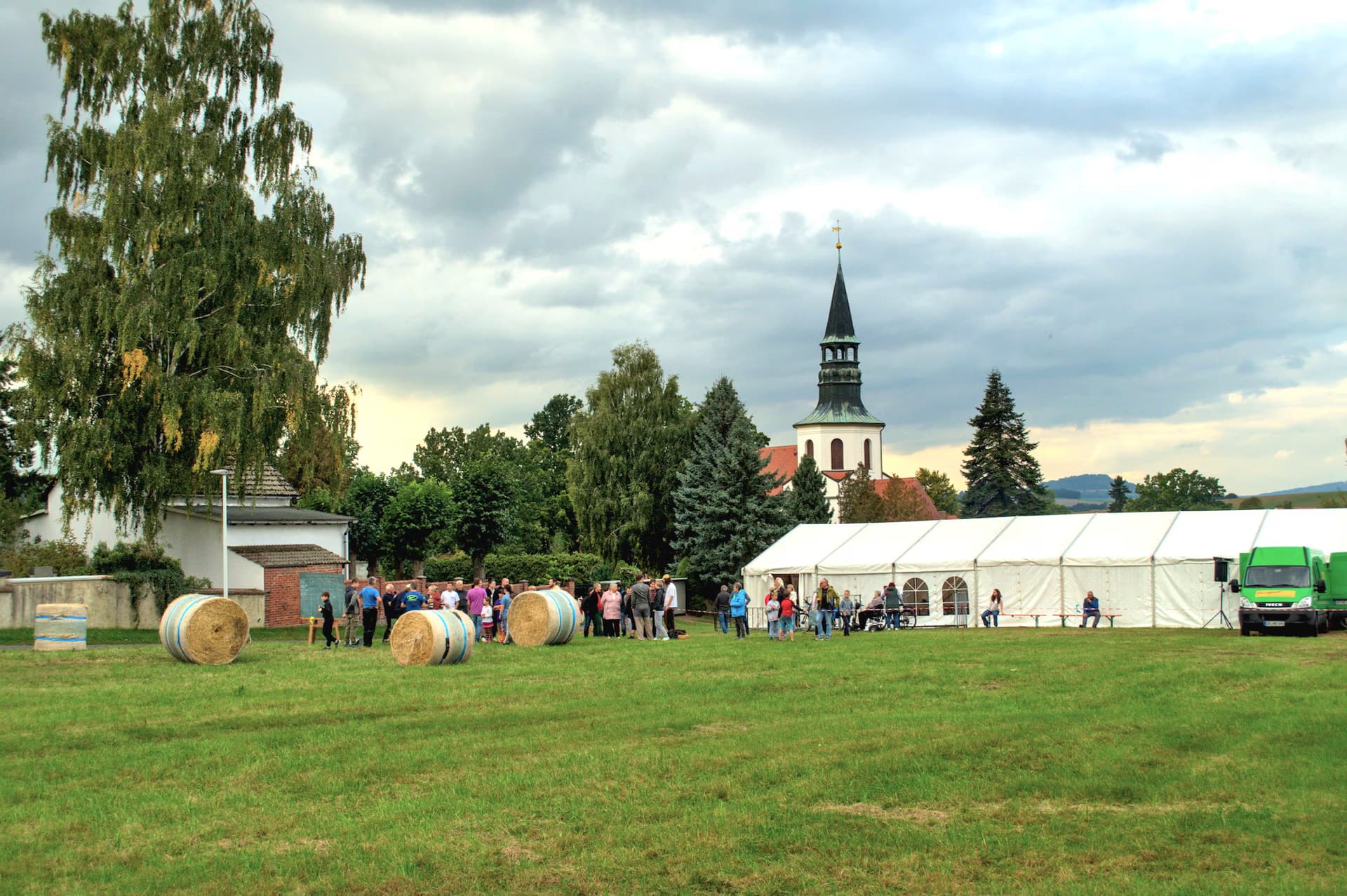 People gathered on a grassy field with hay bales and a large white tent near a church.