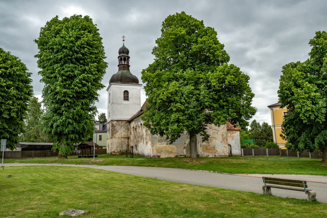 Eine alte Kirche mit verwitterter Steinfassade und einem hohen weißen Turm steht zwischen grünen Bäumen.
