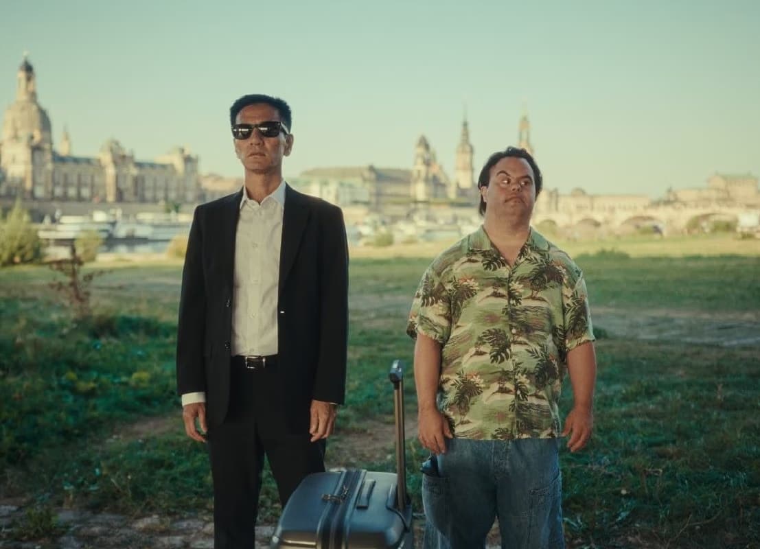 Two men stand outdoors before the Dresden skyline with the Frauenkirche visible in the background.