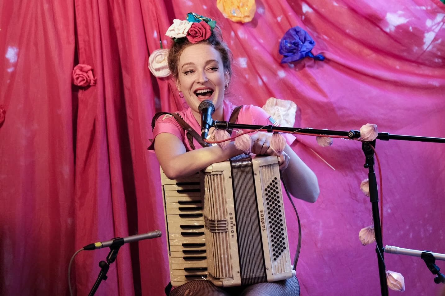 A smiling woman with floral hair ornaments performs while playing an accordion against pink drapes.