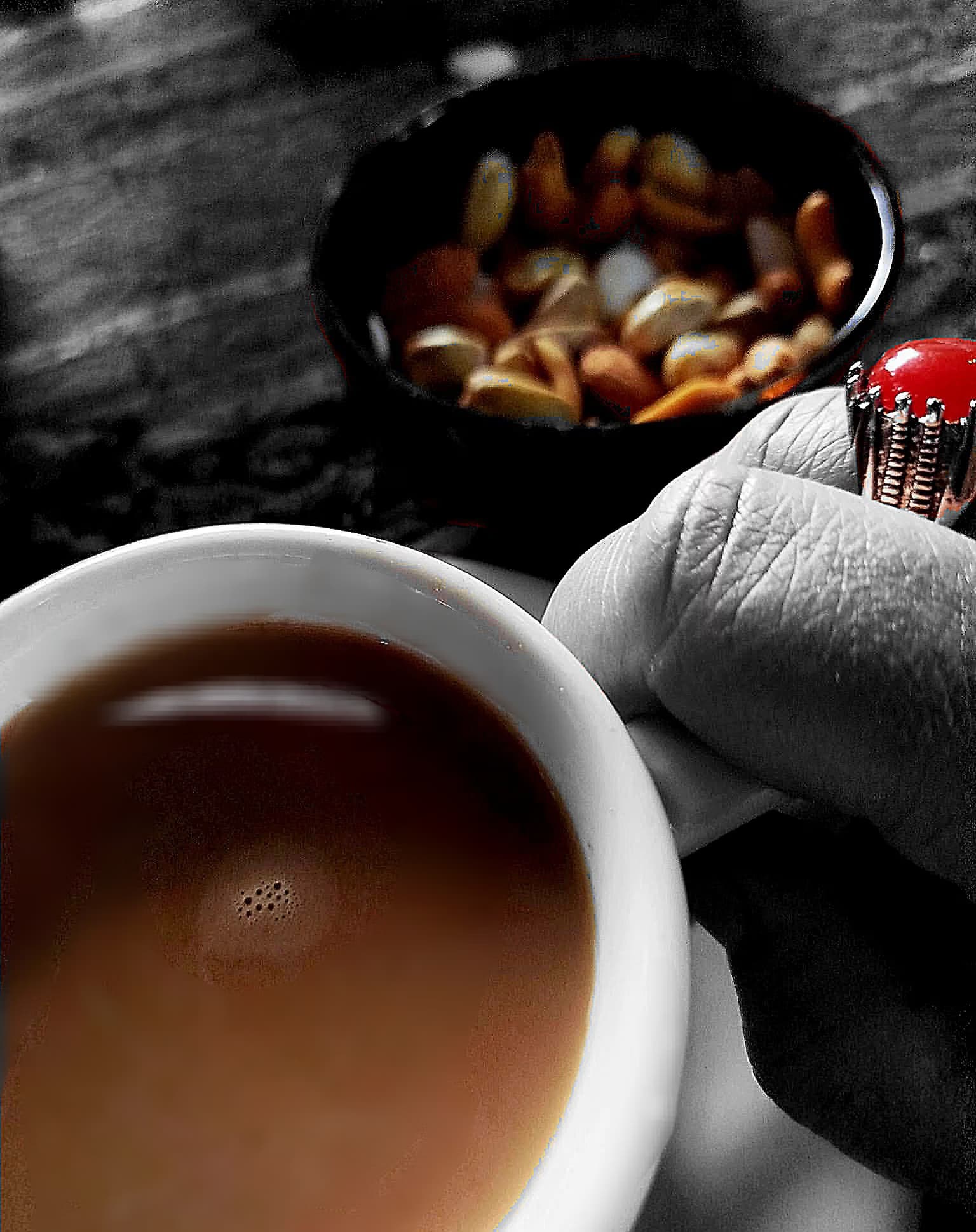 A hand wearing a ring holds a cup of coffee near a bowl of nuts.