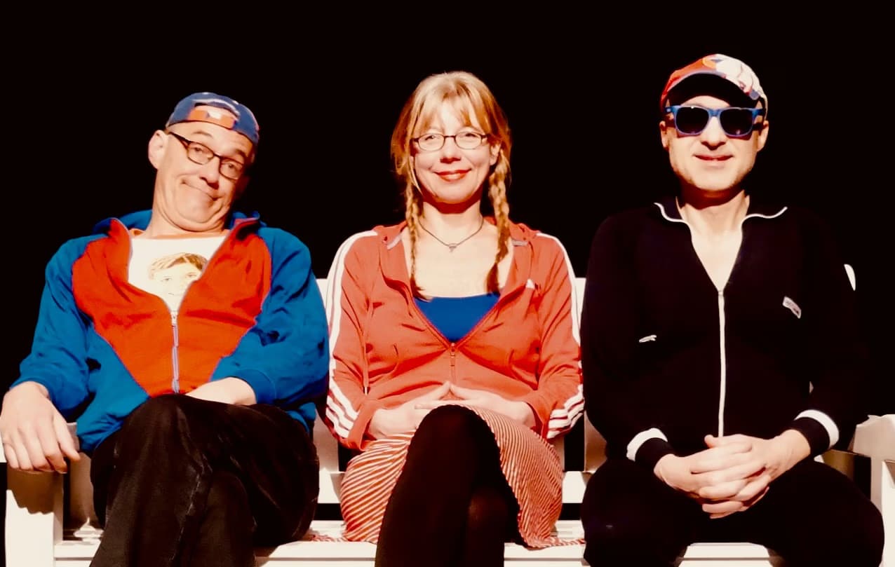 Three people sit on a bench, posing against a black background.