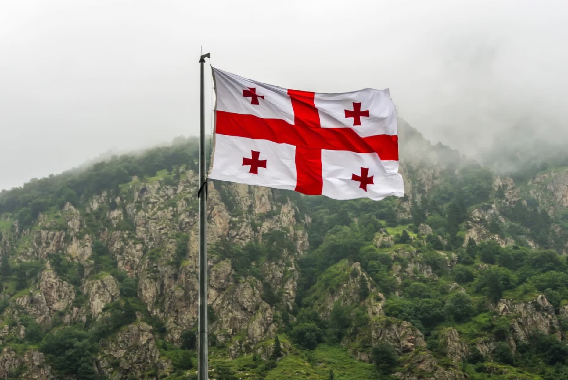 The national flag of Georgia waves on a pole before a foggy mountain landscape.