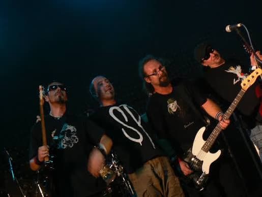 A four-piece rock band performs on a dimly lit concert stage with instruments.