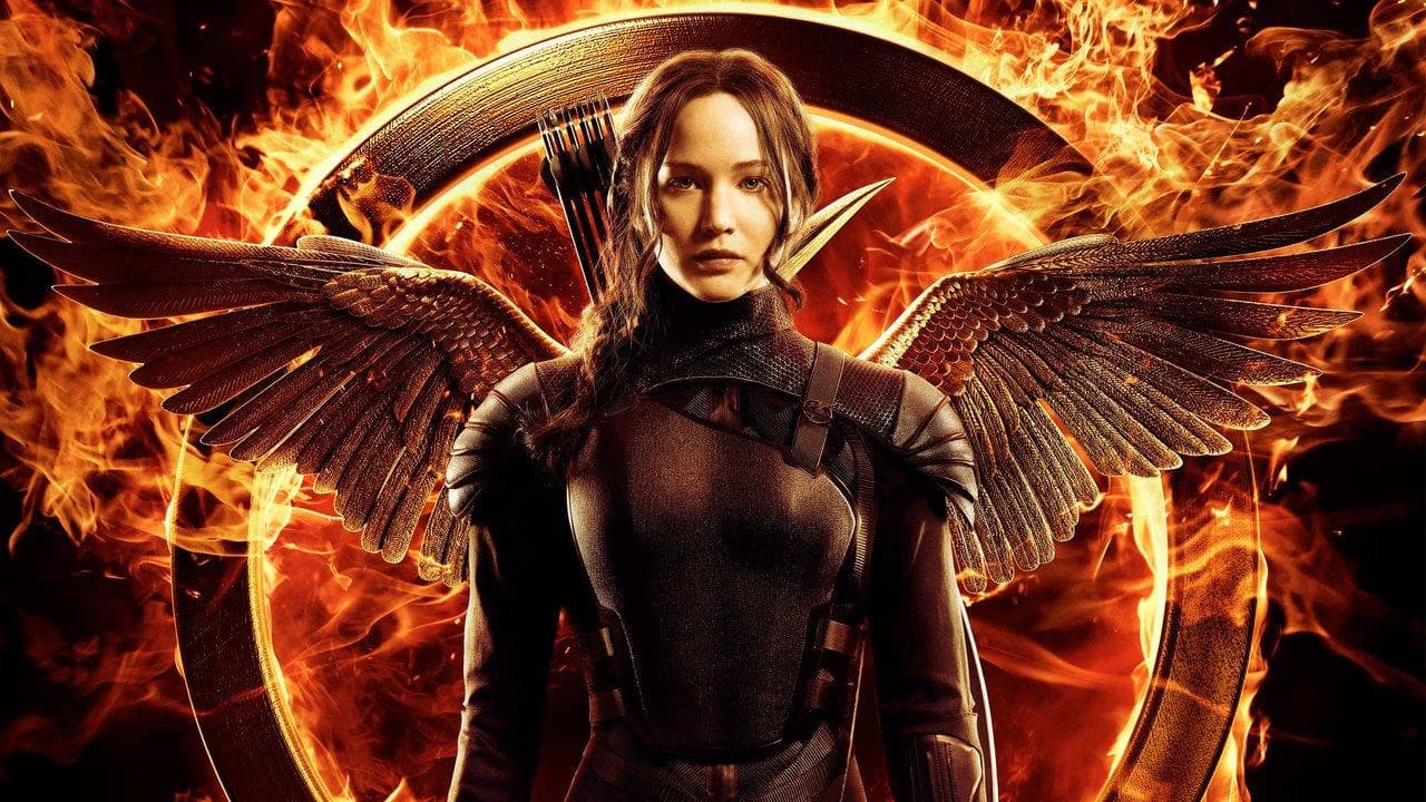 Katniss Everdeen poses with wings against a fiery background and a circular metal emblem.