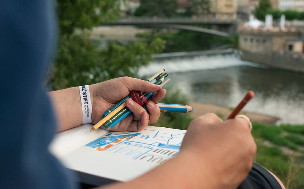 Person sketching outdoors with a view of a river and bridge in the background.