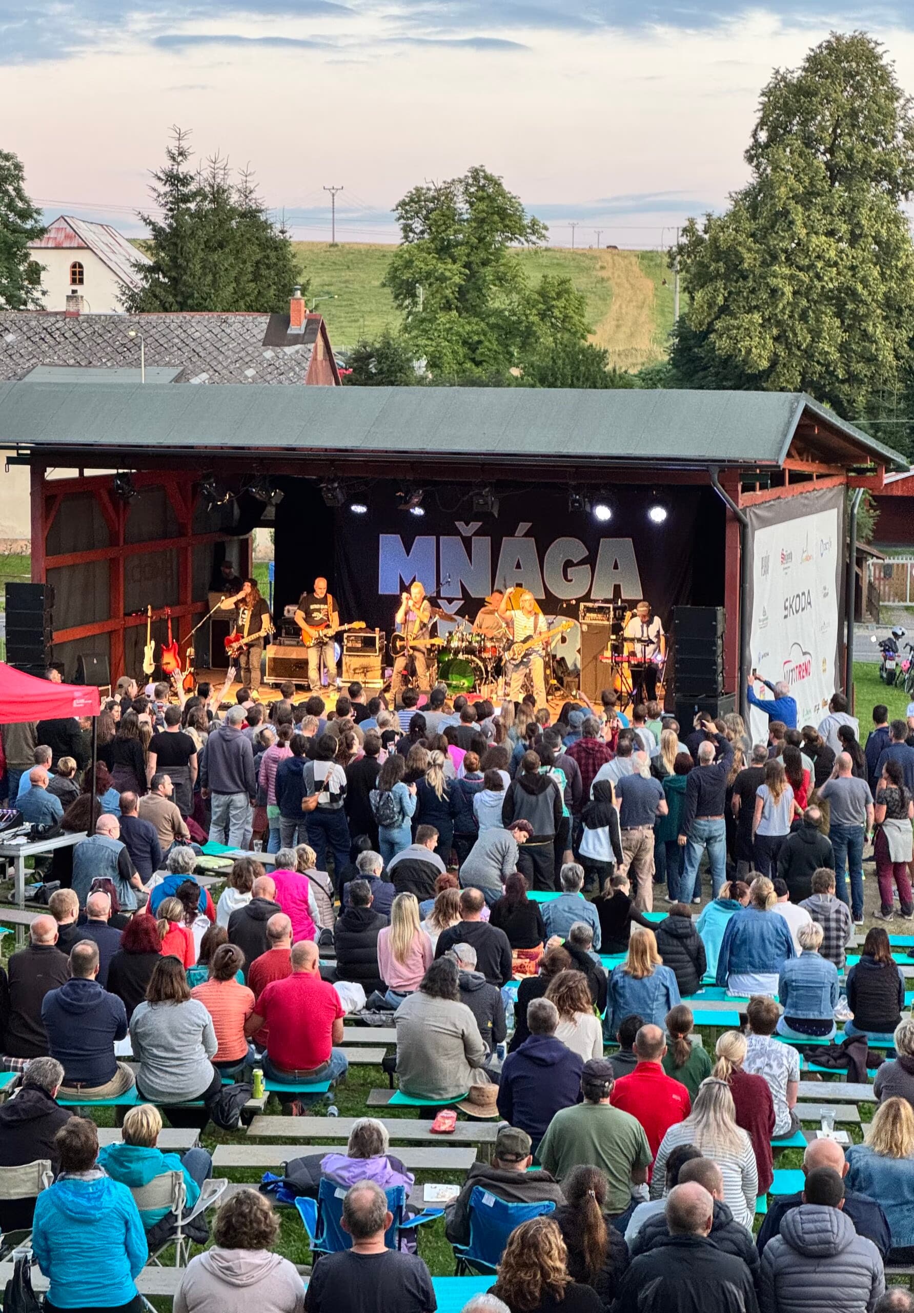 A large crowd sits watching a live band perform on an outdoor wooden stage at dusk.