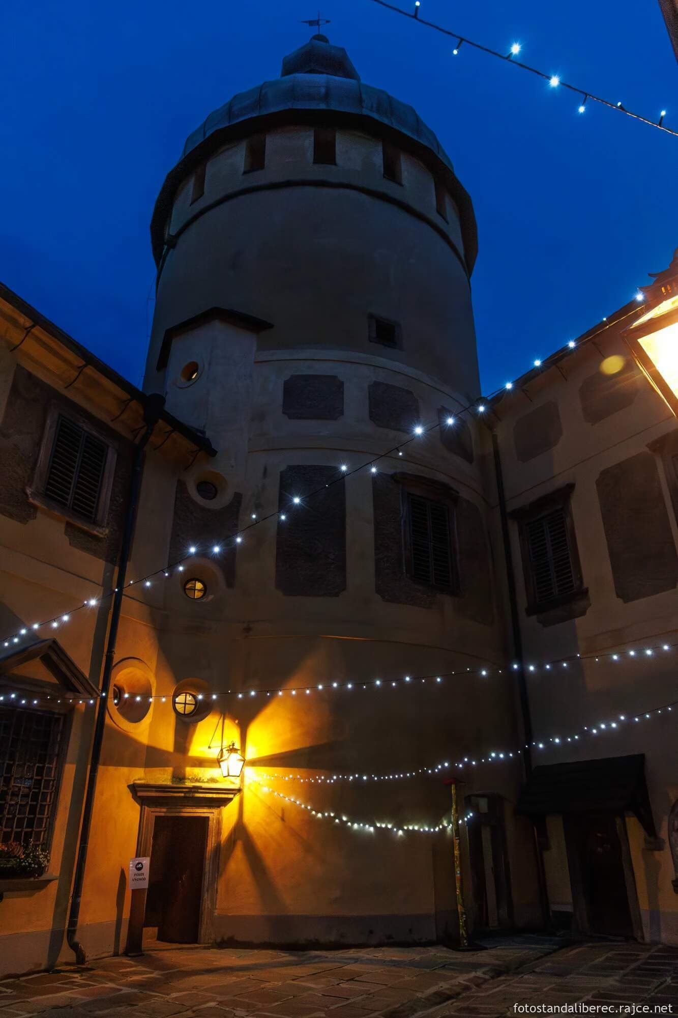A stone tower and courtyard are illuminated by hanging string lights at night.
