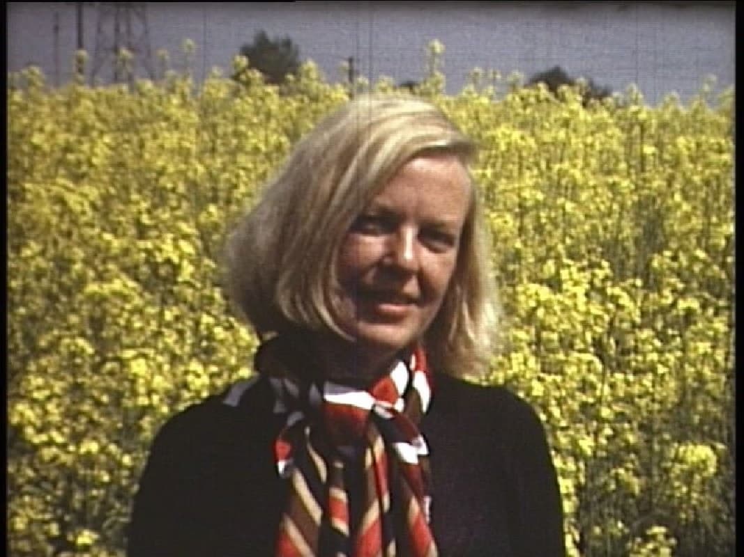 A blonde woman with a patterned scarf smiles in front of a blooming yellow rapeseed field.