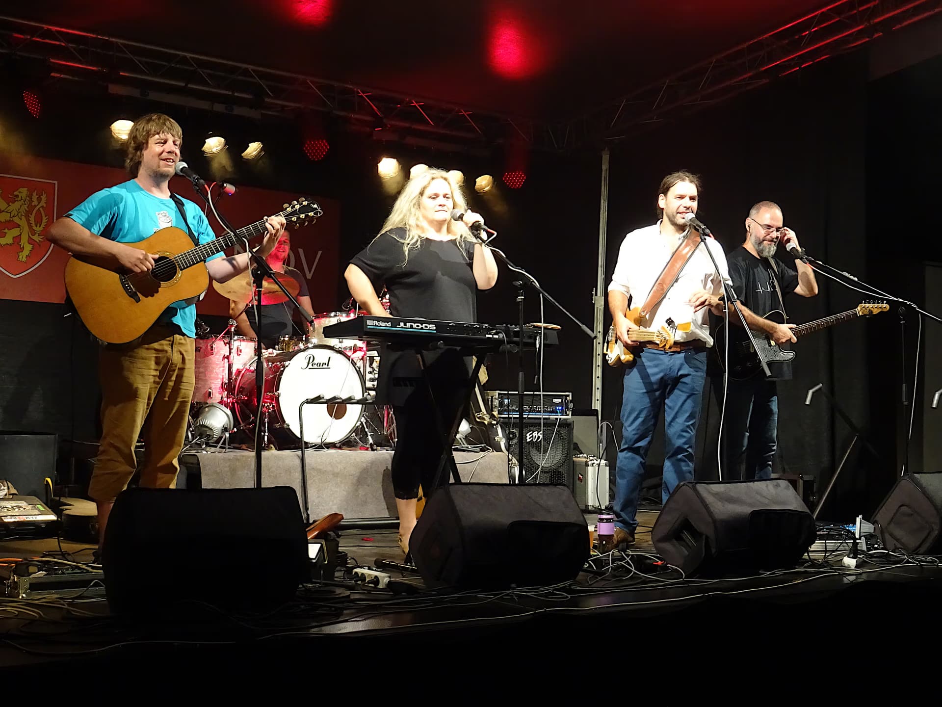 Four band members perform live on stage with instruments and microphones in a dark venue.