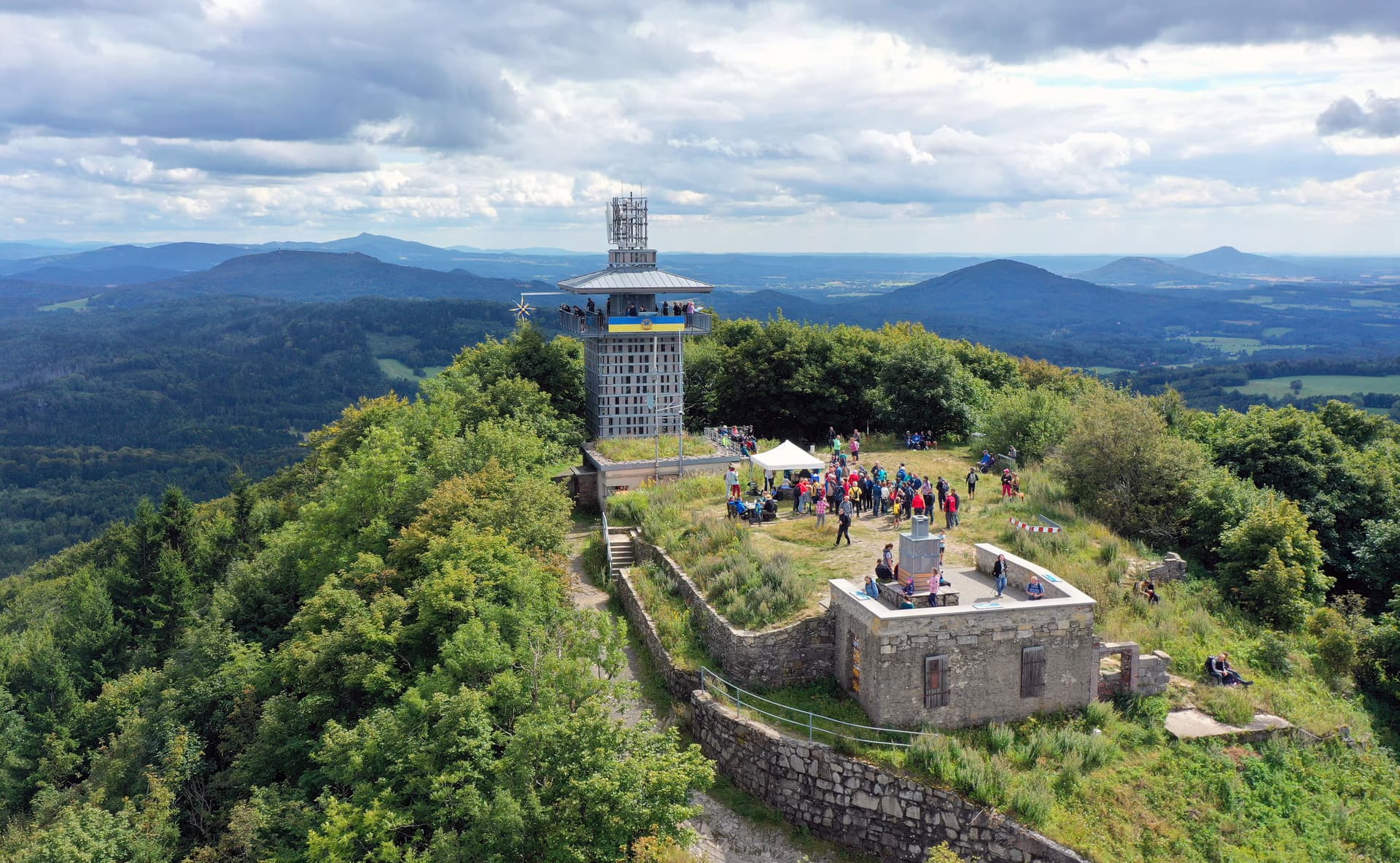 Aerial view of a stone lookout tower on a forested mountain summit with people gathering below.